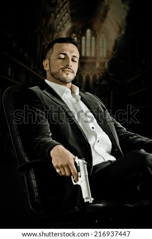 Elegant handsome latin man gangster mafia spy hitman assassin with closed eyes sitting in a chair holding a gun over dark background