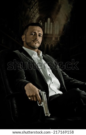 Stylish hispanic young handsome man model mobster spy hitman killer sitting in a chair holding a gun over dark background