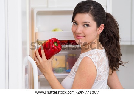 young beautiful brunette woman searching for food in the fridge holding red pepper