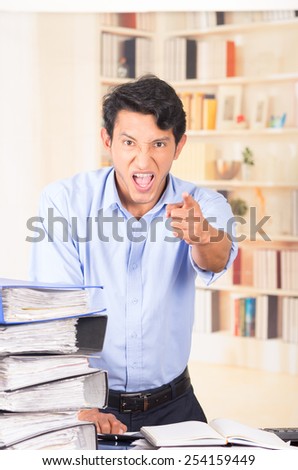 young stressed overwhelmed business man with piles of folders on his desk screaming and pointing at camera