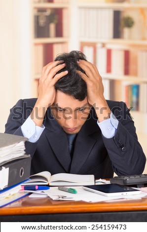 young stressed overwhelmed business man holding head with his hands looking at piles of folders on his desk