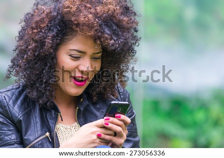 exotic beautiful young girl with dark curly hair using her cell phone texting and sitting in the garden