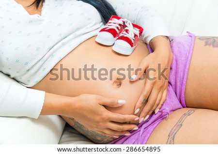 caption of pregnant belly of woman lying sideways with converse baby shoes sitting on stomach plus tattoos visible hands on stomach