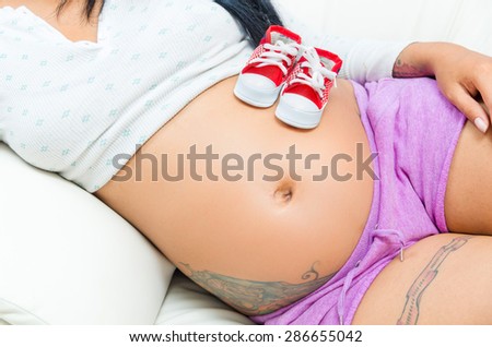 caption of pregnant belly of woman lying sideways with converse baby shoes sitting on stomach plus tattoos visible