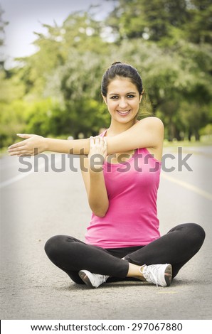 Hispanic brunette in yoga clothing sitting with legs crossed using right arm to stretch the left while smiling at camera.