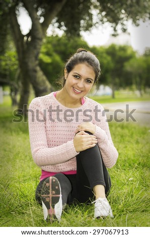 Hispanic brunette sitting on grass in yoga clothing left knee bent with arms wrapped around looking towards camera smiling..