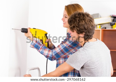 Couple renovating together as woman using power drill on wall with man standing next to her observing.