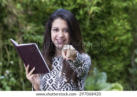 Hispanic brunette in park environment wearing formal clothing holding book open while pointing into camera and smiling.
