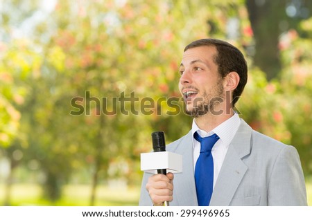 Successful handsome male news reporter in light grey suit working outdoors in park environment holding microphone in live broadcasting.