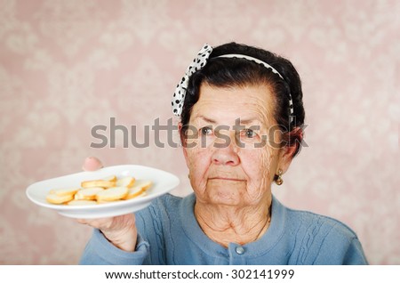Older cute hispanic woman wearing blue sweater and polka dot bowtie on head holding up a plate of cookies.