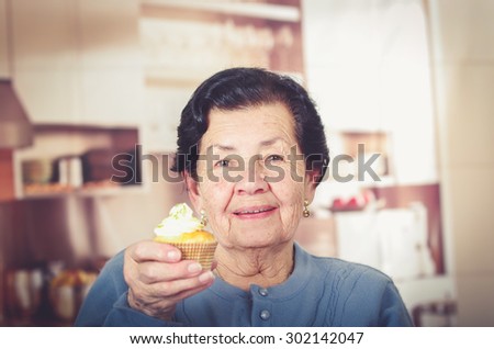 Older hispanic happy woman wearing blue sweater sitting in front of camera showing off cupcake with cream topping.