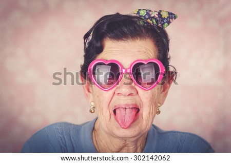 Older cool hispanic woman wearing blue sweater, flower pattern bow on head and pink heartshaped sunglasses looking into camera showing her tongue.
