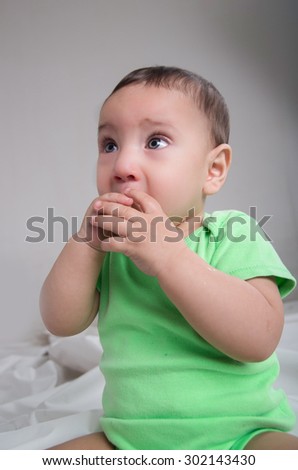 cute baby boy wearing green clothing sitting in front of camera covering mouth with hands and staring to the side.
