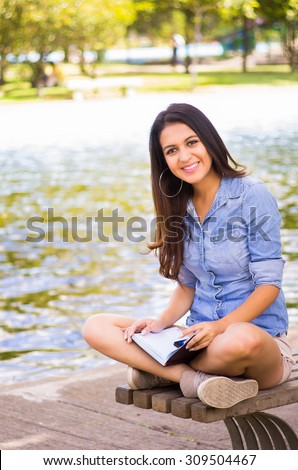 Brunette model wearing denim shirt and white shorts relaxing in park environment, sitting on bench next to lake smiling at camera.