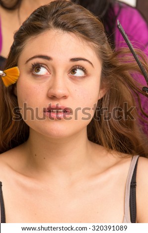 Closeup headshot brunette model facing camera getting makeup done by professional stylist using orange brush.