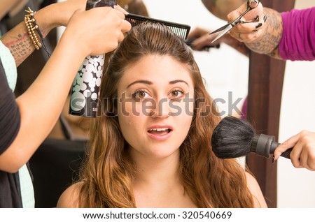 Brunette model facing camera is getting makeup and hair done by two professional stylists as they use different brushes tools.