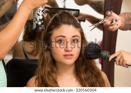 Brunette model facing camera is getting makeup and hair done by two professional stylists as they use different brushes tools.