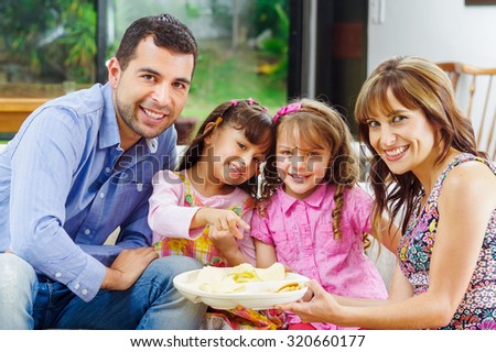 Hispanic parents with two daughters enjoying a tray of potato chips sitting in sofa while smiling and enjoying each other company.
