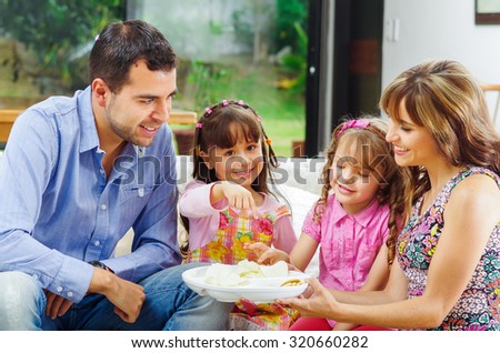 Hispanic parents with two daughters enjoying a tray of potato chips sitting in sofa while smiling and enjoying each other company.
