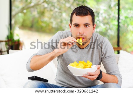 Hispanic male wearing light blue sweater plus denim jeans sitting in white sofa holding bowl of potato chips and remote control watching tv enthusiastically.