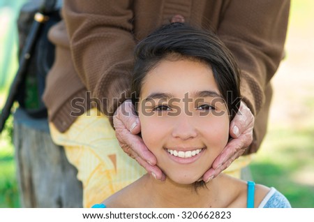 Lovely picture young girl headshot smiling naturally into camera with grandmothers hands holding her head.