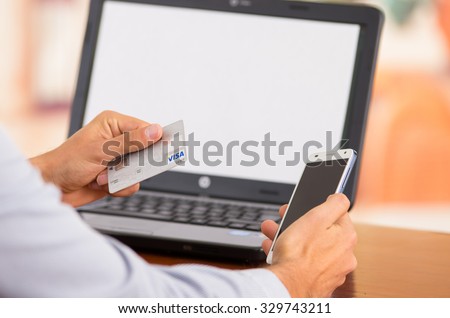 QUITO, ECUADOR - AUGUST 3, 2015: Closeup of young mans hands holding smartphone up, Visa card in other hand with laptop computer sitting on desk.