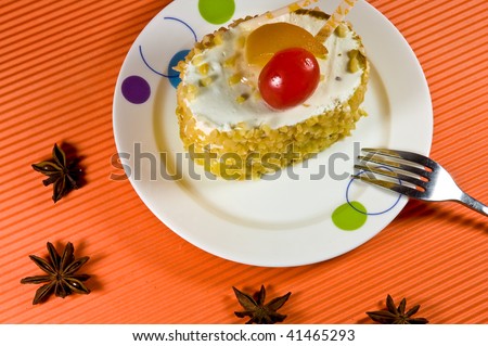 Tasty yellow nut  cake with white cream and red berry. Cake is placed on the white plate along with the shiny metal fork upon the orange background.