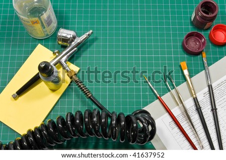 Different painting hobby tools (airbrush, brushes, paints, cutting matt and tweezers) still life composition.