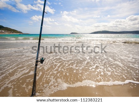 Fixed fishing rod set up on a beach near the surf on the incoming tide.  Blue sky with clouds, turquoise sea.