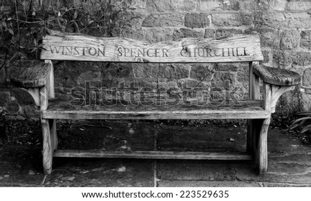 Wooden garden bench with the name Winston Spencer Churchill carved in the back from Chartwell House, Kent, his home until he died in 1965.  Against a stone wall on stone flagging.  Black and white .