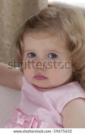 Baby girl,with piercing blue eyes, looking serious,  nearly two years old in a pink dress posing for the camera.