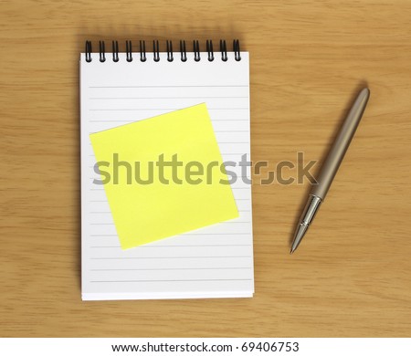 blank yellow sticky note stuck to spiral notebook with pen on wooden desk background. Add your own text