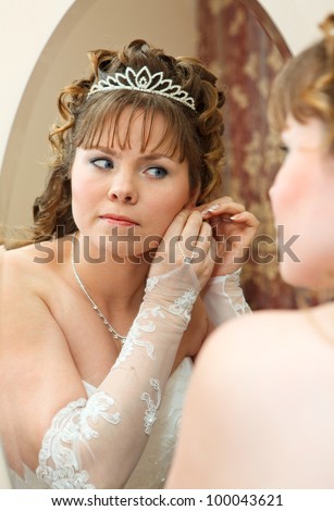Beauty young Caucasian bride with curly hair looking in mirror and wearing earrings