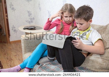 Two Caucasian kids having fun with pad while sitting on sofa in domestic room