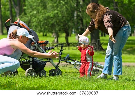 Two women help toddler to do first steps in the park. Green grass and foliage around they. It`s a summer. Baby carriage and bike are here too.