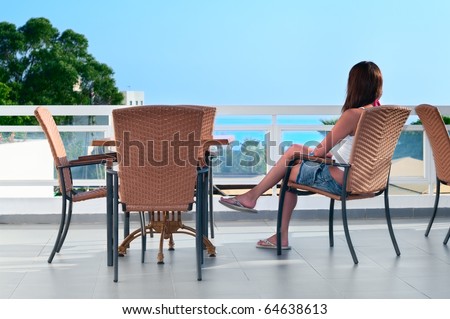Young beautiful girl sitting on a chair on the balcony of the hotel and looks into the distance. One Caucasian woman is back side