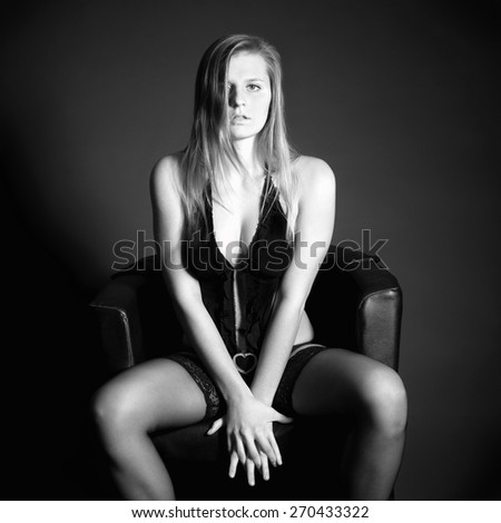 Black and white fashion portrait of a beautiful woman in erotic lingerie sitting on black armchair in front of dark studio background