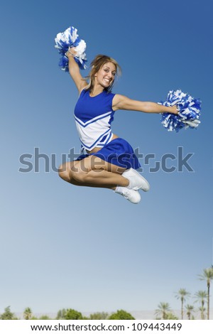 Full length of a young cheerleader jumping with pom-poms against sky