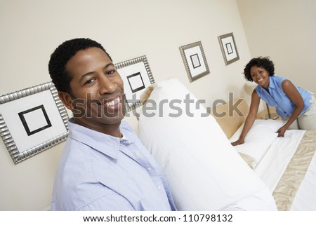 Portrait of an African American man holding pillow with woman arranging bed at home
