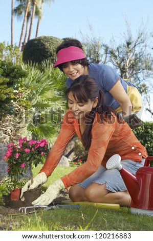 Happy daughter planting flower plant with mother standing in the background at garden