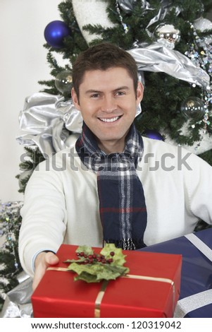Portrait of a happy young man giving gift box with Christmas tree in the background