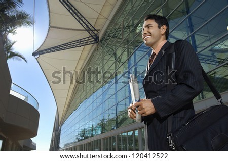 Happy businessman with shoulder bag and newspaper in front of office building