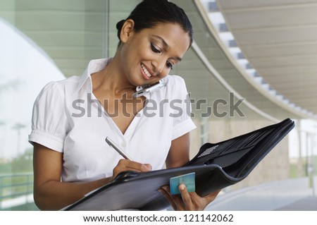 An Indian business woman communicating on mobile phone while writing notes in the folder