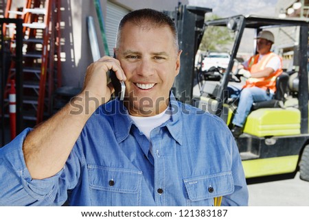 Portrait of a happy male industrial worker communicating on cell phone with his coworker in the background