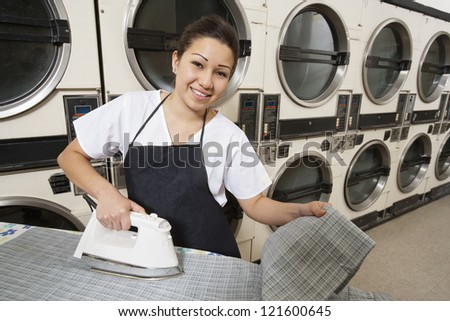 Portrait of a happy woman wearing apron ironing in front of washing machines