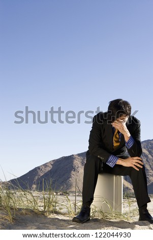 Full length of a depressed businessman sitting on briefcase in desert