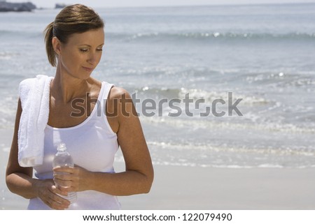 Sporty woman holding water bottle with towel on shoulder at beach