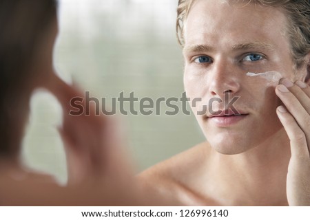 Mirror reflection of a young man applying facial cream on face