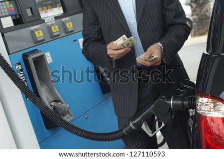 Mid section of businessman counting money with gasoline refueling car at fuel station