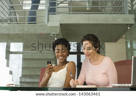 Young businesswoman using cellphone with female colleague noting down on notepad in office